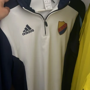 Vit och marinblå Adidas DIF långärmad tränings tröja - Snygg vit och marinblå vindjacka från Adidas med Djurgårdens IF-logga på bröstet. Jackan har halv dragkedja, lång ärm och sportig look. Perfekt för dig som vill representera DIF med stil. Materialet är polyester och jackan har en clean, modern design.