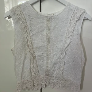 Vit spetsblus från ONLY, stl 36 - Superfin vit blus från ONLY med broderade blommor och spetsdetaljer längs kanterna. Ärmlös modell med knappar i ryggen och vackert hålmönster framtill. Perfekt för dig som gillar romantisk och söt stil.