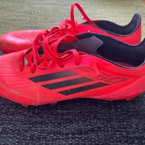 Röda fotbollsskor från Adidas PRO  - Snygga röda fotbollsskor från Adidas med svarta detaljer och klassiska tre ränder på sidan. Skorna har snörning, låg profil och är tillverkade i syntetmaterial för lätt känsla på planen. Perfekta för dig som vill sticka ut på matchen.