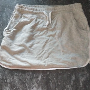 Grå sportig kortkjol med snörning - Sportig ljusgrå kortkjol i mjukt bomullsmaterial med vita kanter och snörning i midjan. Kjolen har två praktiska fickor framtill och en avslappnad passform, perfekt för en chill och aktiv stil.