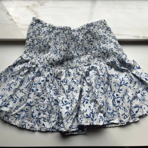 Vit och blå blommig kjol från H&M - Supersöt kort kjol från H&M i vitt med blått blommönster. Kjolen har smockad midja som sitter skönt och volangig nederdel för en luftig känsla. Kjolen är knappast använd.