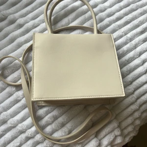 Vit axelväska  - Snygg beige axelväska från H&M med stilren, fyrkantig form. Väskan har både handtag och lång axelrem så du kan bära den på flera sätt. Tillverkad i slätt syntetmaterial och har ett rymligt fack inuti. Perfekt för att lyfta vilken outfit som helst.