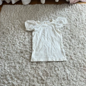 Vit topp med puffärmar i spets - Supersöt vit topp med korta puffärmar i transparent spets och rund halsringning. Perfekt för dig som gillar romantiska detaljer och en feminin look. Toppen är figurnära och har en enkel, stilren design med extra fokus på ärmarna.