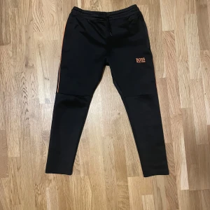 Svarta mjukisbyxor Hugo Boss M - Svarta mjukisbyxor från Hugo Boss med orangea detaljer längs sidorna och logga på låret. Byxorna har elastisk midja med snörning och är tillverkade i mjukt material. Perfekta för chill eller träning, med smal passform och diskreta fickor. Storlek M känns som S.