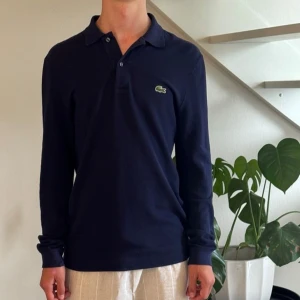 Mörkblå långärmad piké från Lacoste - Säljer en mörkblå långärmad pikétröja från Lacoste i slim fit. Tröjan har klassisk krage, knappar vid halsen och den ikoniska gröna krokodilloggan på bröstet. Tillverkad i bomull för en skön och stilren känsla.
