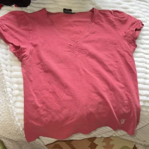 Rosa v-ringad tshirt - Rosa topp från Jonna B. med v-ringning och korta ärmar. Toppen har en liten glittrig krona broderad nertill och rynkade detaljer vid halsringningen. Perfekt för dig som gillar enkla men snygga detaljer.  Mer mauve-färgad än på bilden! 