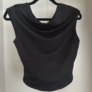 Svart croppad ärmlös topp - Snygg svart croppad topp med draperad halsringning och ärmlös design. Perfekt för dig som gillar minimalistisk och stilren look.