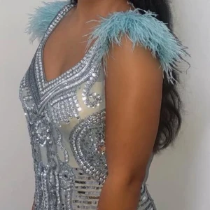Glittrig silverklänning med fjädrar - Säljer en glittrig silverfärgad klänning med v-ringning och ljusblå fjädrar på axlarna. Klänningen har massor av paljetter och pärldetaljer som ger en lyxig känsla. Perfekt för dig som vill sticka ut och glänsa på festen!