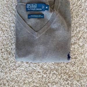 Grå v-ringad tröja Polo Ralph Lauren - Grå v-ringad tröja från Polo Ralph Lauren i 100% bomull. Klassisk passform med långa ärmar och broderad marinblå logga på bröstet. Perfekt för dig som gillar stilrena och tidlösa plagg med en clean look.