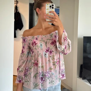 Blommig offshoulder blus  - Supersöt blommig offshoulder blus från Amisu i ljusrosa med lila, gröna och vita blommor. Smockad upptill och vid ärmslut, vida trekvartsärmar och flowy passform. Perfekt för en somrig och romantisk look.