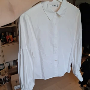 Vit skjorta från NA-KD, stl 36 - Klassisk vit skjorta från NA-KD i storlek 36. Skjortan har långärmade ärmar med breda manschetter och knappar framtill. Rak passform och stilren krage, perfekt till jeans eller kjol. Ballongärms känsla. Vissa små mörka fläckar se sista bild, påverkar ej helheten.