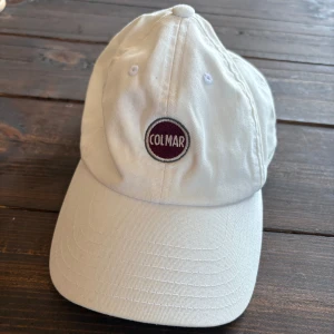 Vit keps från Colmar - Snygg vit keps från Colmar med klassisk böjd skärm och justerbart spänne bak. Framtill finns en rund Colmar-logga i vinrött och vitt. Kepsen är tillverkad i bomull och har ventilerande broderade hål för extra komfort.