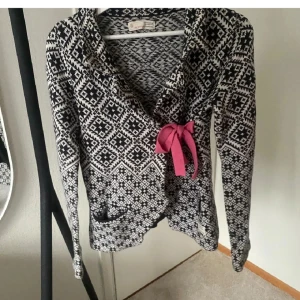 Odd Molly Kofta  - Supersnygg svartvit kofta från Odd Molly med stickat mönster och flera rosa rosetter som detaljer. Koftan har omlottknäppning med en rosa rosett framtill, långa ärmar och två små fickor med rosetter bak. Storlek S/M.