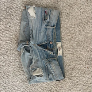 Ljusa jeansshorts från Hollister - Snygga ljusblå jeansshorts från Hollister med slitna detaljer och råa kanter. Klassisk femficksmodell med låg midja och broderad logga på fickan. Perfekta för varma dagar och en avslappnad stil.