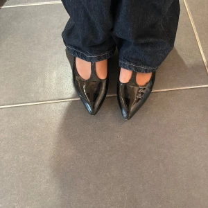 Svarta ballerinaskor Bianco - Snygga svarta ballerinaskor från Bianco i glansigt lackmaterial. Skorna har spetsig tå, T-rem och smal ankelrem med spänne. Perfekta för dig som vill ha en stilren och trendig look med lite edge.