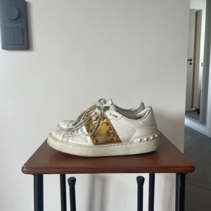 Valentino Garavani sneakers - Snygga vita Valentino Garavani Rockstud sneakers med guldiga detaljer och nitar på sidorna. Skorna har snörning och en chunky sula med ikoniska pyramidnitar runt hälen. Tillverkade i skinn och har en cool, lyxig vibe. Dem är nogorlunda slitna men främst inuti skon, det är inget som känns eller syns när man har dem på sig! 