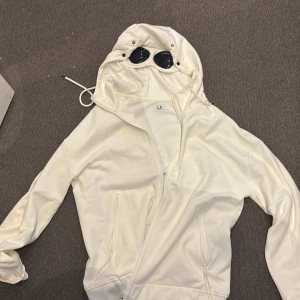 Vit C.P. Company goggle jacka XL - Säljer en vit goggle jacka från C.P. Company i storlek XL. Jackan har huva med ikoniska svarta glasögon, dragkedja framtill och två fickor med dragkedja. Materialet är mjukt och har använt 1 gång så är i otroligt bra skick