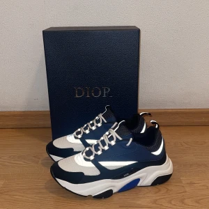 Chunky sneakers från Dior B22 i blått och vitt - Snygga chunky sneakers från Dior i marinblått, vitt och svart med inslag av reflekterande detaljer. Skorna har en kraftig sula, meshpartier och vita snören. Perfekta för dig som vill sticka ut med exklusiv fin stil