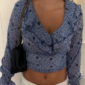 Blå mönstrad croppad blus - Säljer en blå croppad blus med lång ärm och coolt grafiskt mönster. Blusen har v-ringning och volangdetalj längs urringningen och axlarna. Tunt, lätt material som känns luftigt och fräscht. Perfekt för dig som gillar statement-plagg med snygg passform.