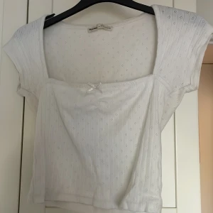 Vit blus med rosett från Pull&Bear - Supersöt vit blus från Pull&Bear med små broderade hålmönster och en söt liten rosett framtill. Toppen har korta ärmar, fyrkantig urringning och croppad passform. Perfekt för en fräsch och romantisk look till sommaren.