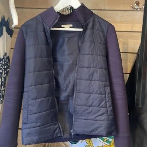 Mörklila pufferjacka från Angel & Rocket - Snygg mörklila pufferjacka från Angel & Rocket med quiltad framsida och släta ärmar. Jackan har dragkedja och hög krage, perfekt för kyliga dagar. Materialmix med polyester och syntet ger en modern look. Enkel och stilren design som passar till det mesta.