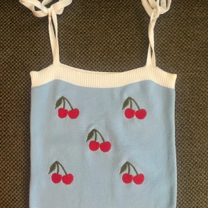 Ljusblå stickad linnetopp med körsbär - Supersöt ljusblå stickad linnetopp med vita ribbade kanter och knytbara axelband. Toppen har broderade röda körsbärsmotiv över hela framsidan. Perfekt för en somrig look och passar till både jeans och kjol.
