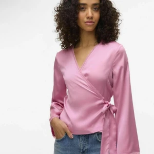 Rosa omlottopp med knytband - Snygg rosa omlottopp med v-ringning och knyt i sidan. Toppens material har en lätt glans och känns mjukt mot huden. Långa ärmar och diskret spetskant längs ringningen ger en extra touch. Perfekt att styla med jeans eller kjol. Storlek M men sitter mer som S