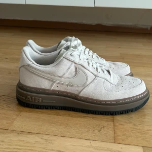 Nike Air Force 1 LUXE - Snygga Nike Air Force 1 sneakers i beige mocka med grov brun sula och klassisk snörning. Skorna har perforerade detaljer på tån och en ikonisk Swoosh-logga på sidan. Perfekta för dig som gillar stilrena och trendiga sneakers med en twist.