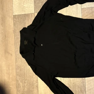 Svart merinoull pikétröja Massimo Dutti XL - Klassisk svart långärmad pikétröja från Massimo Dutti i mjuk merinoull. Tröjan har krage och en knapp framtill, perfekt för en clean och stilren look. Passar dig som gillar minimalistiskt och tidlöst mode.