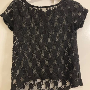 Svart spetsblus med dragkedja - Snygg svart spetsblus med korta ärmar och en dekorativ dragkedja i ryggen. Perfekt för en stilren look med sitt genombrutna mönster. Tillverkad i 100% polyester.