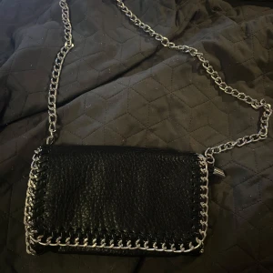 Svart axelväska med kedja från Tiamo - Säljer en svart axelväska från Tiamo i skinn med grov silverfärgad kedja som axelrem. Väskan har snygga detaljer med kedja runt kanterna som är avtagbar.