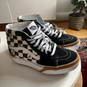Vans Sk8-Hi checkerboard sneakers - Snygga Vans Sk8-Hi sneakers med klassiskt svart och vitt checkerboard-mönster, vita snören och detaljer i mocka. Skorna har hög modell, vit sula med brun rand och ikonisk Vans-logga baktill. Perfekta för dig som gillar streetstyle och skatekänsla.