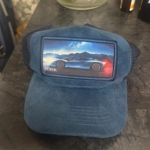 Blå keps med bilmotiv från PERIK - Snygg blå keps med mesh-bakstycke och justerbar snapback. Framsidan har en cool patch med sportbilsmotiv mot berg och himmel. Kepsen har mockaliknande tyg framtill och klassisk böjd skärm. Perfekt för dig som gillar bilar och streetstyle.