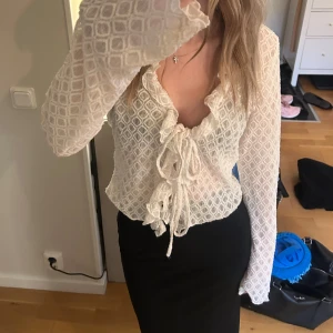 Vit omlottblus med snörning - Supersnygg vit blus med omlott och snörning framtill. Blusen har ett unikt rutmönster och långa ärmar. Materialet är lätt och luftigt, perfekt för vår och sommar. Den djupa v-ringningen och volangdetaljerna ger en trendig vibe.