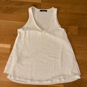 Vit linnetopp från Mango - Fräsch vit linnetopp från Mango med djup v-ringning och ärmlös design. Toppen har en lätt, luftig känsla och är tillverkad i ett mjukt bomullsmaterial. Perfekt för varma dagar och enkel att matcha med jeans eller shorts.