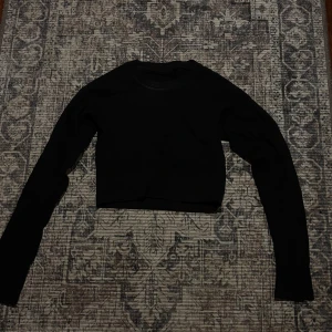 Svart croppad långärmad topp XS/S - Svart croppad långärmad topp från SOSPOT, tillverkad i Italien. Materialet är mjukt och stretchigt med 92% polyamid och 4% elastan, vilket gör den riktigt skön och följsam. Enkel och clean design med rund hals och tight passform.