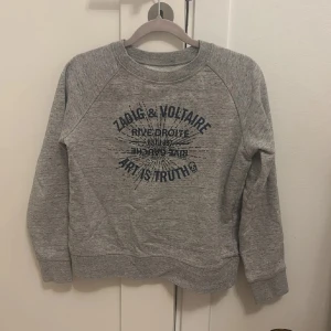 Grå sweatshirt från Zadig & Voltaire - Säljer en grå sweatshirt från Zadig & Voltaire med mörkblå tryck på bröstet där det står 'Art is Truth'. Tröjan har rund hals, långa ärmar och ribbade muddar. Perfekt för dig som gillar streetstyle och vill ha något bekvämt och snyggt.