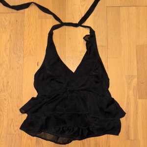 Svart omlott topp från Nelly - Säljer en svart omlott topp från Nelly med volangdetaljer och knyt i nacken. Toppen är ärmlös och har en djup v-ringning samt ett lätt, luftigt tyg som ger en snygg flowy look. Perfekt för dig som gillar trendiga och stilrena plagg.