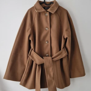 Brun kappa med bälte från Gina Tricot - Snygg brun kappa från Gina Tricot i storlek XS. Jackan har klassisk krage, knäppning med knappar framtill och ett brett bälte i midjan. 