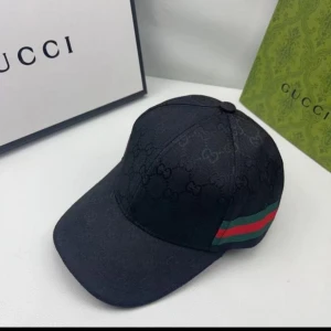 Svart Gucci keps med GG-mönster - Snygg svart keps från Gucci med diskret GG-monogram över hela ytan. Klassisk böjd skärm och justerbar rem bak med metallspänne. På sidan finns Guccis ikoniska gröna och röda ränder. Materialet är slitstark bomull och polyester.