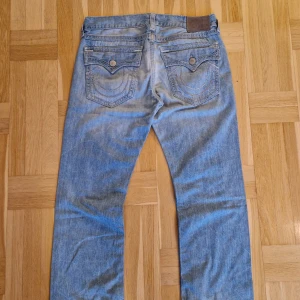 True Religion ljusblå jeans - Säljer ett par klassiska ljusblå jeans från True Religion med snygga kontrastsömmar och ikoniska bakfickor med lock och knappar. Jeansen har en lätt bootcut struktur med en baggy liknande fit.  De är tillverkade i bomull och är perfekta för dig som gillar streetwear, och vill sticka ut lite extra.