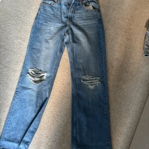 Bershka snygga jeans (aldrig använda) - Säljer ett par blå 90s wide jeans från Bershka i storlek 36. Jeansen har bred passform, klassisk femficksdesign och slitna detaljer över båda knäna för en cool, avslappnad look. Tillverkade i bomull med dragkedja och knapp framtill.
