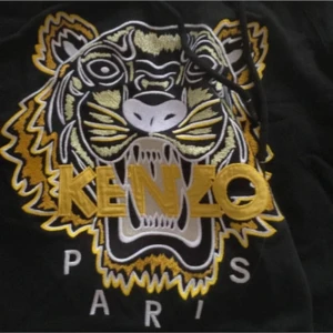 Svart Kenzo hoodie med tigertryck - Svart hoodie från Kenzo med broderat tigerhuvud i gult, vitt och svart på bröstet samt stor Kenzo-logga. Tröjan har huva med snörning och känguruficka framtill. 