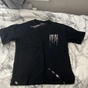 Svart Amiri t-shirt med färgstänk - Svart t-shirt från Amiri med stort tryck på ryggen och mindre tryck på bröstet. Trycket består av AMIRI-loggan med färgstänk i blått, vitt, rött och orange som rinner ner. Klassisk rund hals och korta ärmar. Materialet känns mjukt och är troligtvis bomull.