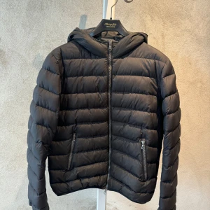 Svart Colmar pufferjacka med huva, storlek 16Y 172 - Svart pufferjacka från Colmar med huva och två dragkedjeförsedda fickor. Jackan har ett quiltat mönster och klassisk logga på ärmen. Perfekt för kyliga dagar och riktigt skön att bära. Stängs med dragkedja hela vägen upp.