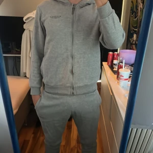 Grå tracksuit byxa och hoodie från McKenzie med dragkedja och luva  - Säljer en grå tracksuit byxa och hoodie från McKenzie med huva och hel dragkedja framtill. Tröjan har lång ärm och är gjord i mjuk bomullsmix med fleece på insidan. Diskret logga på bröstet och ribbade muddar. Perfekt för chill eller träning.