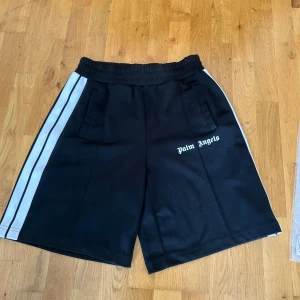 Palm Angels Track Shorts - Svarta track shorts från Palm Angels med vita ränder längs sidorna och logotyp på vänster ben. Elastisk midja och två fickor framtill. Snygg loose passform och sportig vibe, perfekta för chill eller träning. Materialet är polyester som ger en skön känsla. Nypris på dessa 3500