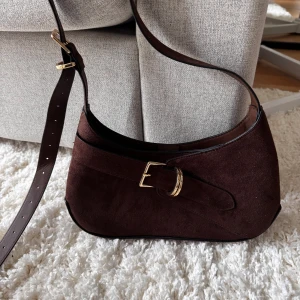 Brun mocka axelväska med spänne - Snygg brun axelväska i mocka med justerbart axelband och guldfärgat spänne framtill. Väskan har en mjuk, rundad form och stängs med magnetlås. Insidan har ett fack med dragkedja för småsaker. Perfekt för dig som gillar stilrena detaljer.