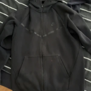 Svart Nike hoodie med dragkedja - Svart hoodie från Nike med hel dragkedja och huva. Klassisk design med diskret Nike-logga på bröstet och två stora fickor framtill. Perfekt för chill dagar eller när du vill ha en sportig look.