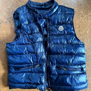 Mörkblå dunväst från Moncler - Snygg mörkblå dunväst från Moncler med glansig finish och klassisk logga på bröstet. Västen har hög krage, två dragkedjefickor och stängs med dragkedja framtill. 